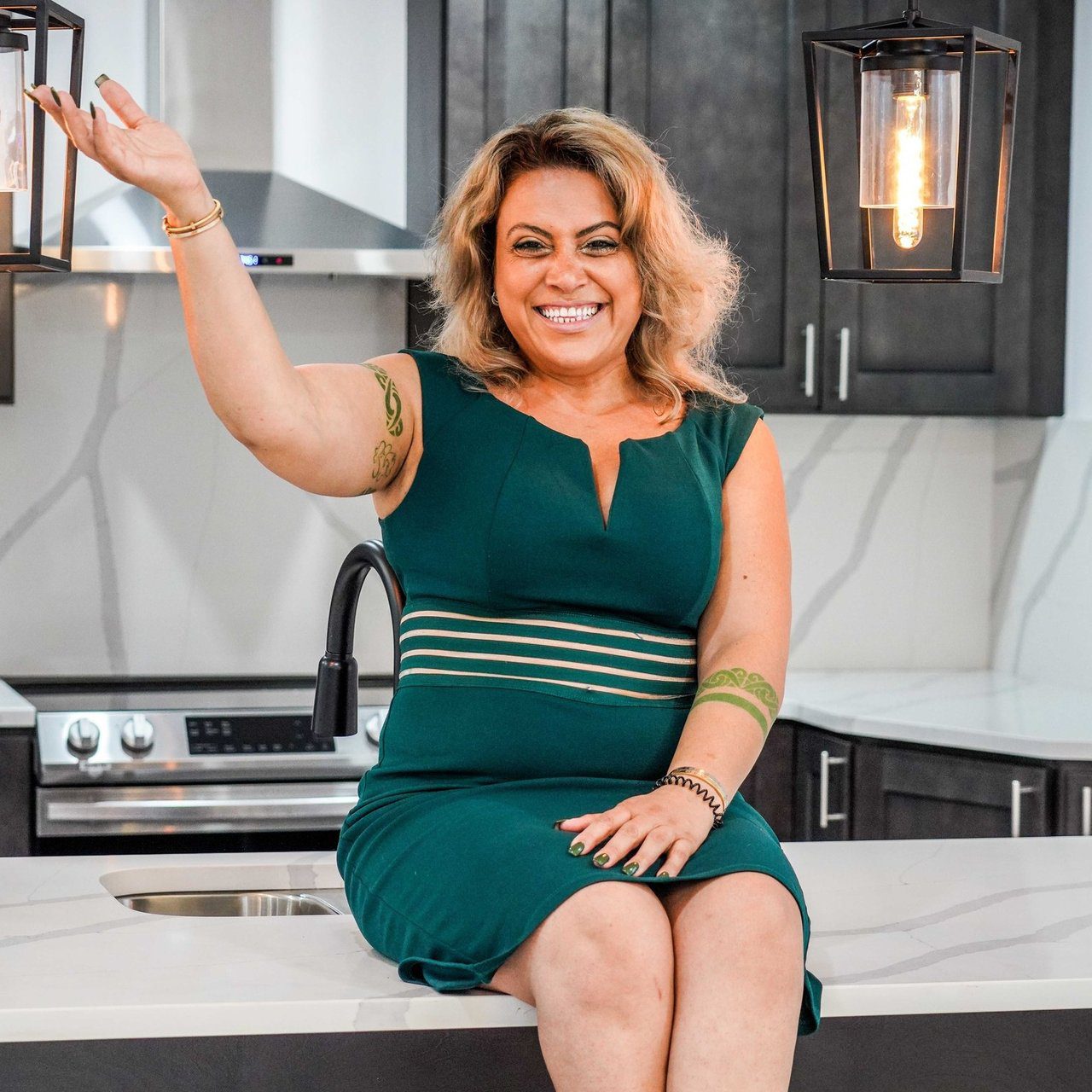 Confident woman in a green dress sitting on a kitchen counter, smiling and gesturing.
