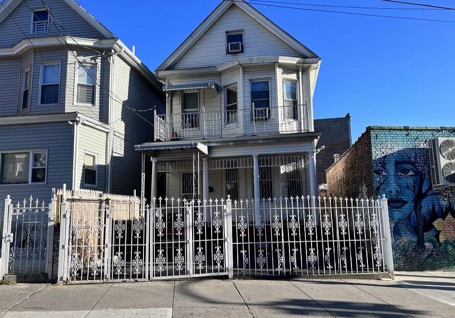 Old two-story house with white exterior and a gated front yard under a clear blue sky.
