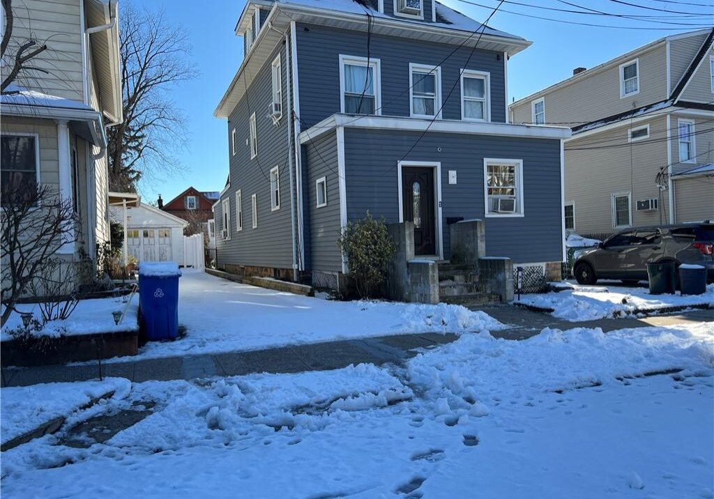 Snow-covered street with a gray house and clear blue sky.